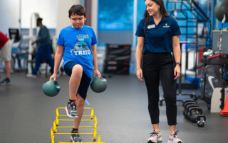 A young boy hold 2 kettlebells as he walks over low hurdles and a physical therapist monitors his balance.