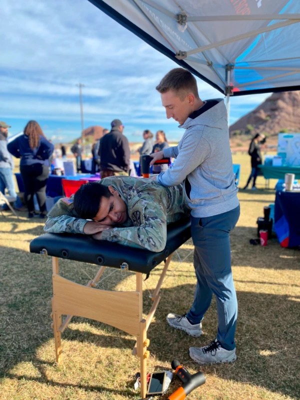 A therapist using a theragun on an army recruit outdoors.