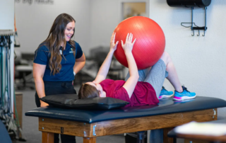 A woman on a table holding a red exercise ball between her hands and knees.