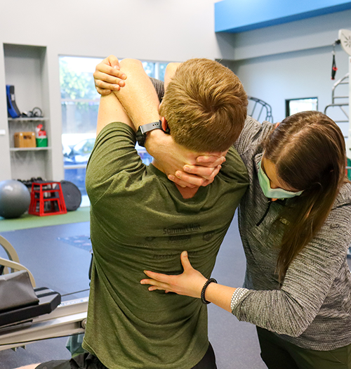 A teen with hands clasp behind his head an a therapist performs a back adjustment.