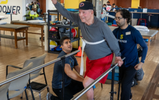 An older man using parallel bars while Valentine Munoz Sanchez and a PT tech help.